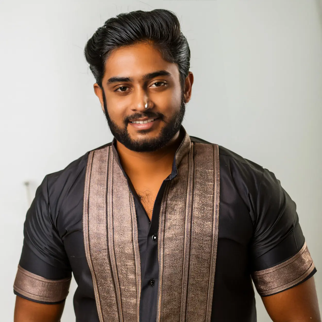 "Young South Asian man wearing black shirt with bronze traditional vest smiling at camera in professional photoshoot"