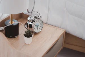 "Minimalist bedside table with silver alarm clock, small succulent plant, and modern lamp on wooden surface"