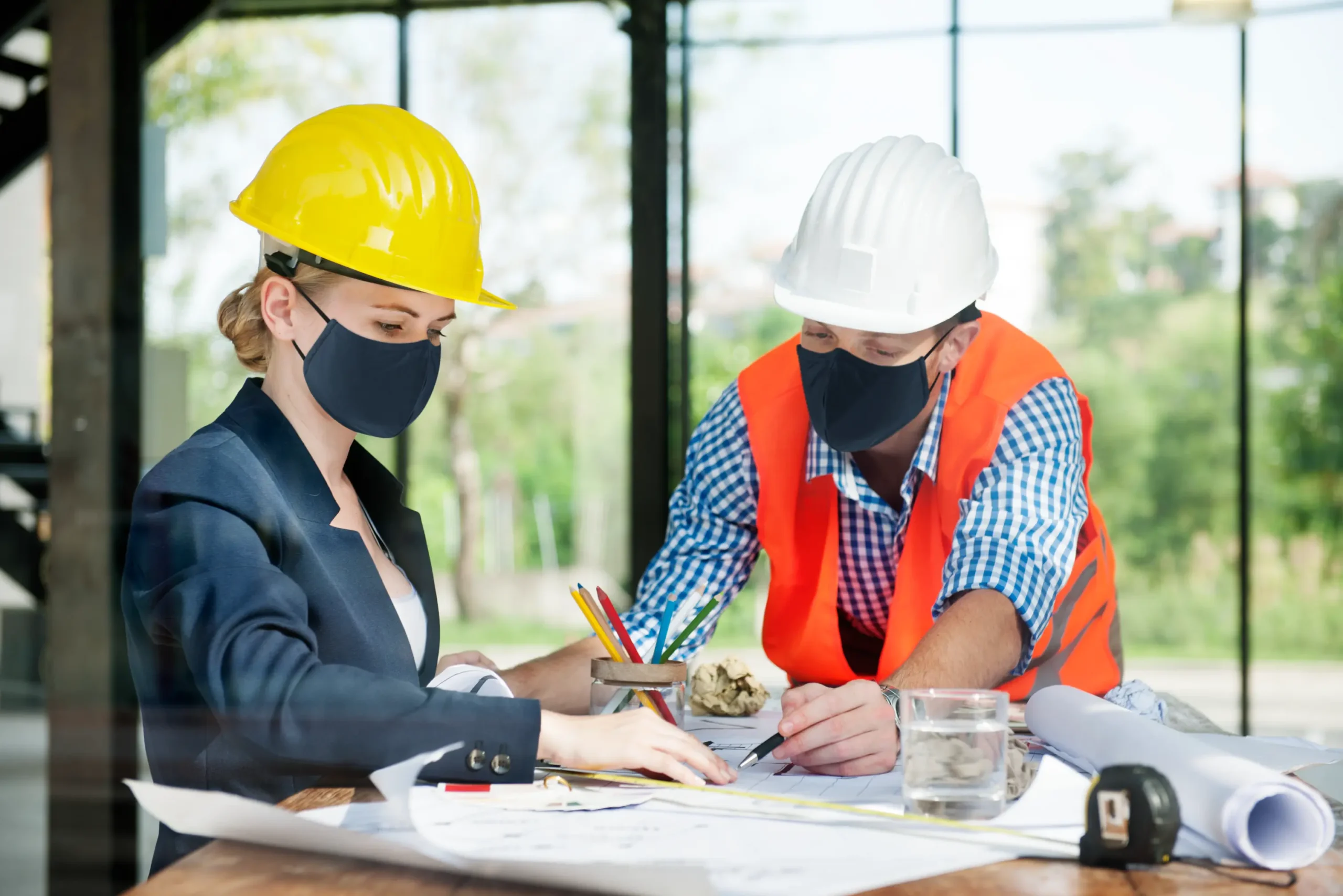 "Construction professionals wearing masks reviewing building plans with safety helmets during pandemic"