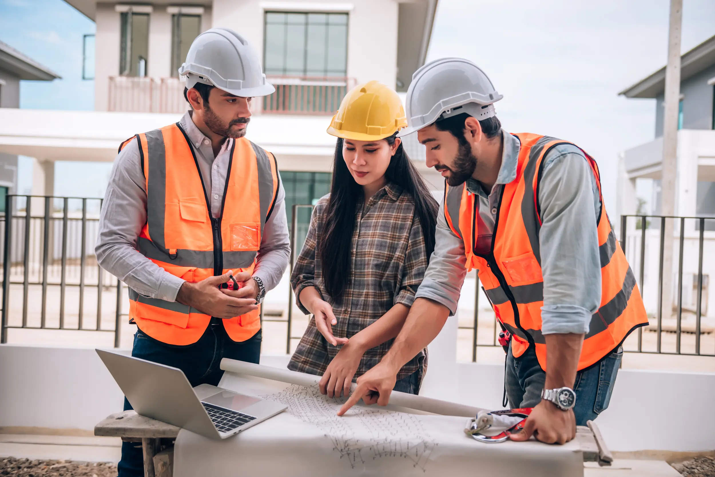 "Construction professionals reviewing building plans with laptop at construction site wearing safety gear and high-visibility vests"