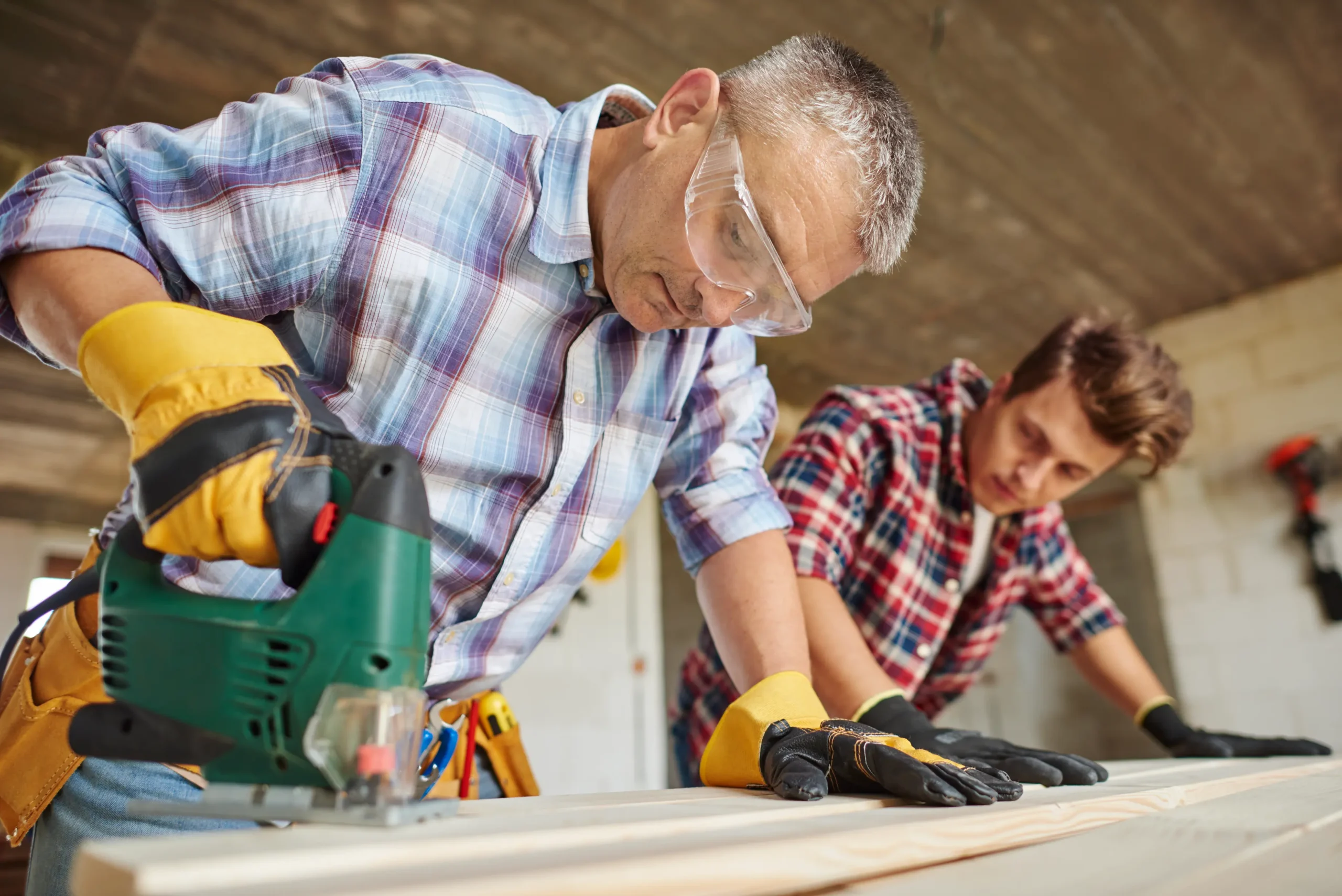 "Experienced carpenter and apprentice working with power tools on wooden surface wearing safety equipment"