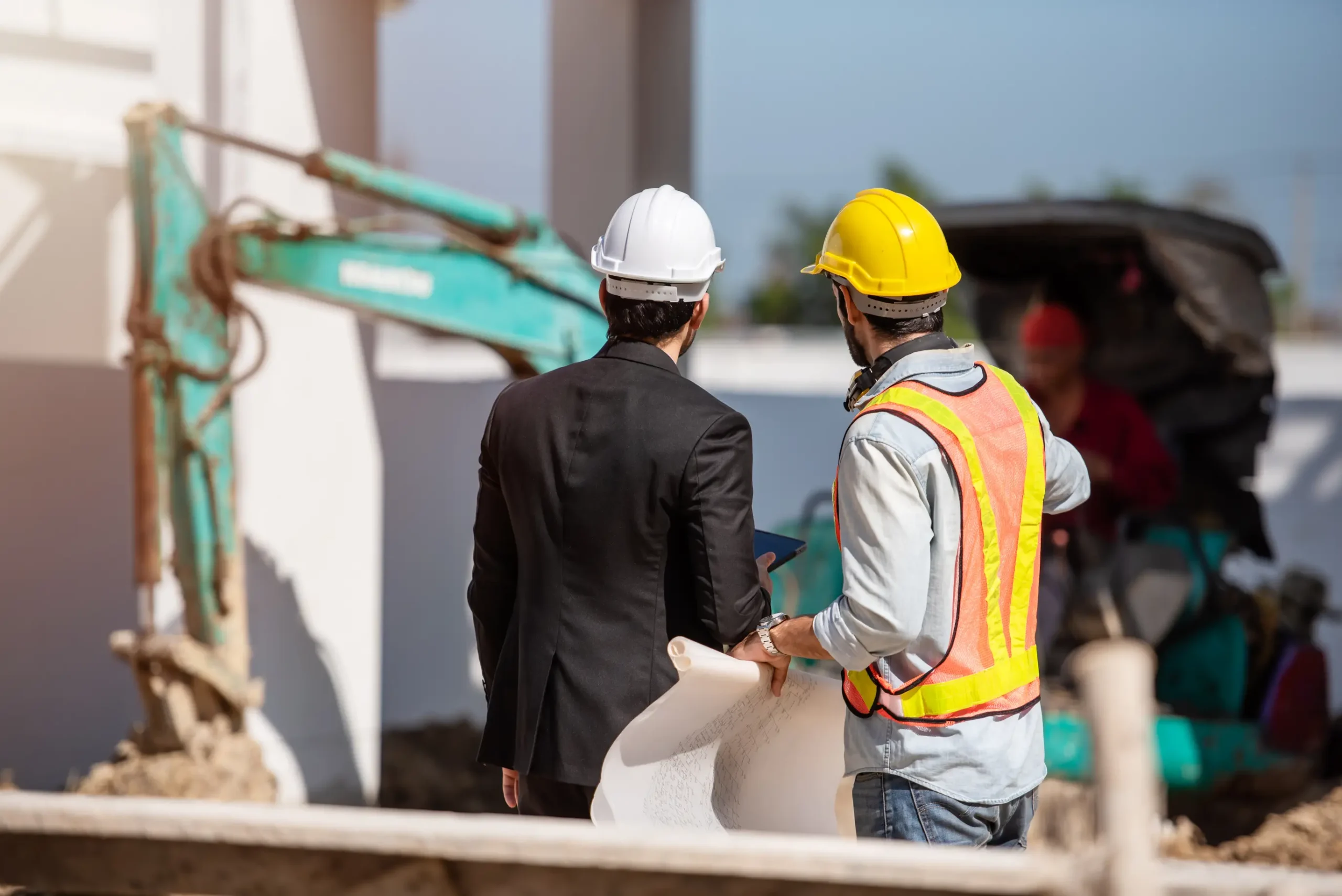 "Construction engineer and site worker reviewing blueprints at construction site with excavator in background"