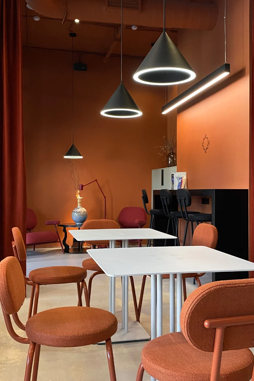 "Modern cafe interior with orange walls, pendant LED lights, white tables and rust-colored upholstered chairs"