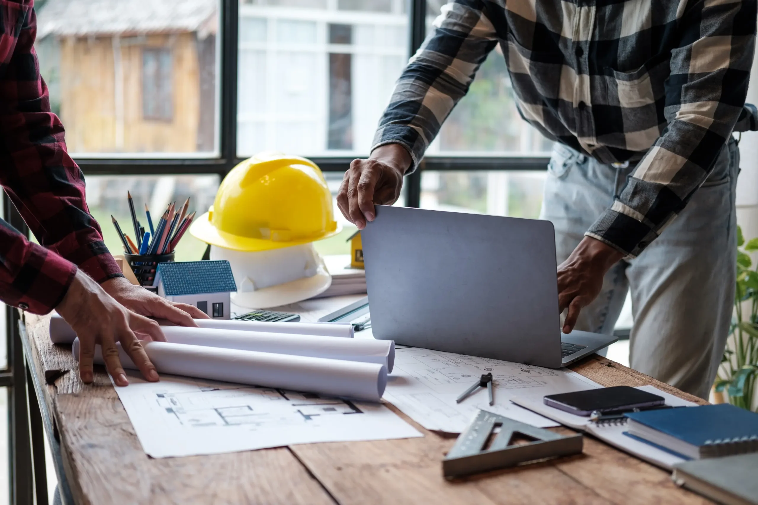 "Construction engineers reviewing architectural blueprints with laptop and yellow hard hat on wooden desk"