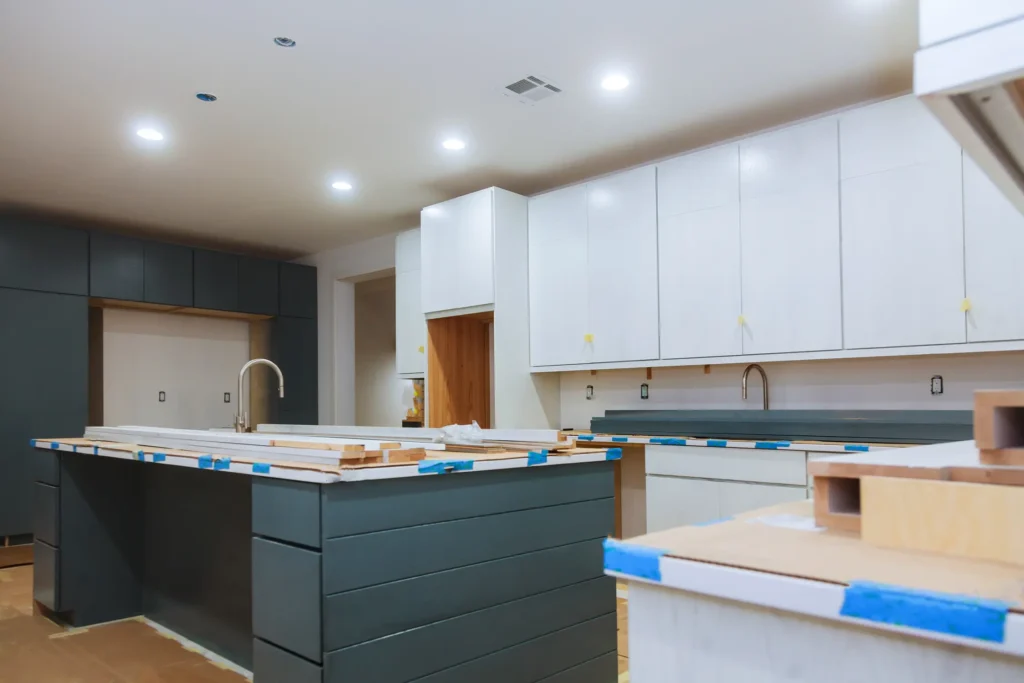 "Modern kitchen renovation in progress with white upper cabinets and dark green island featuring unfinished countertops and painter's tape"