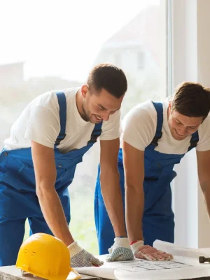 "Construction workers in blue overalls reviewing building blueprints with yellow hardhat on workbench"