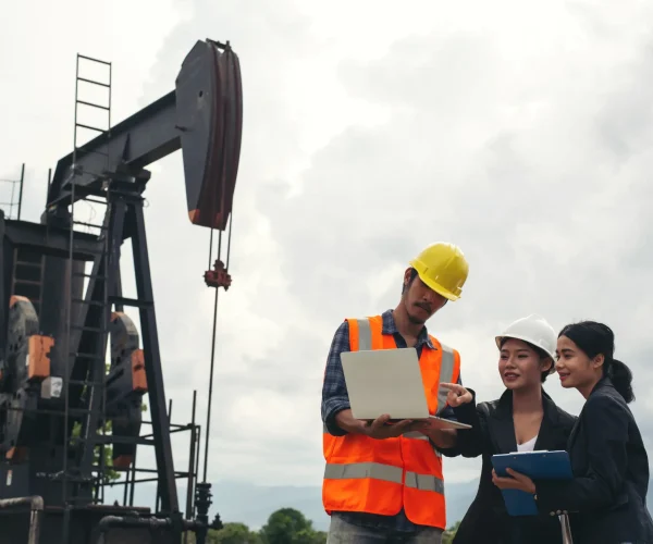 "Petroleum engineers reviewing data on laptop near active oil pump jack in industrial field"